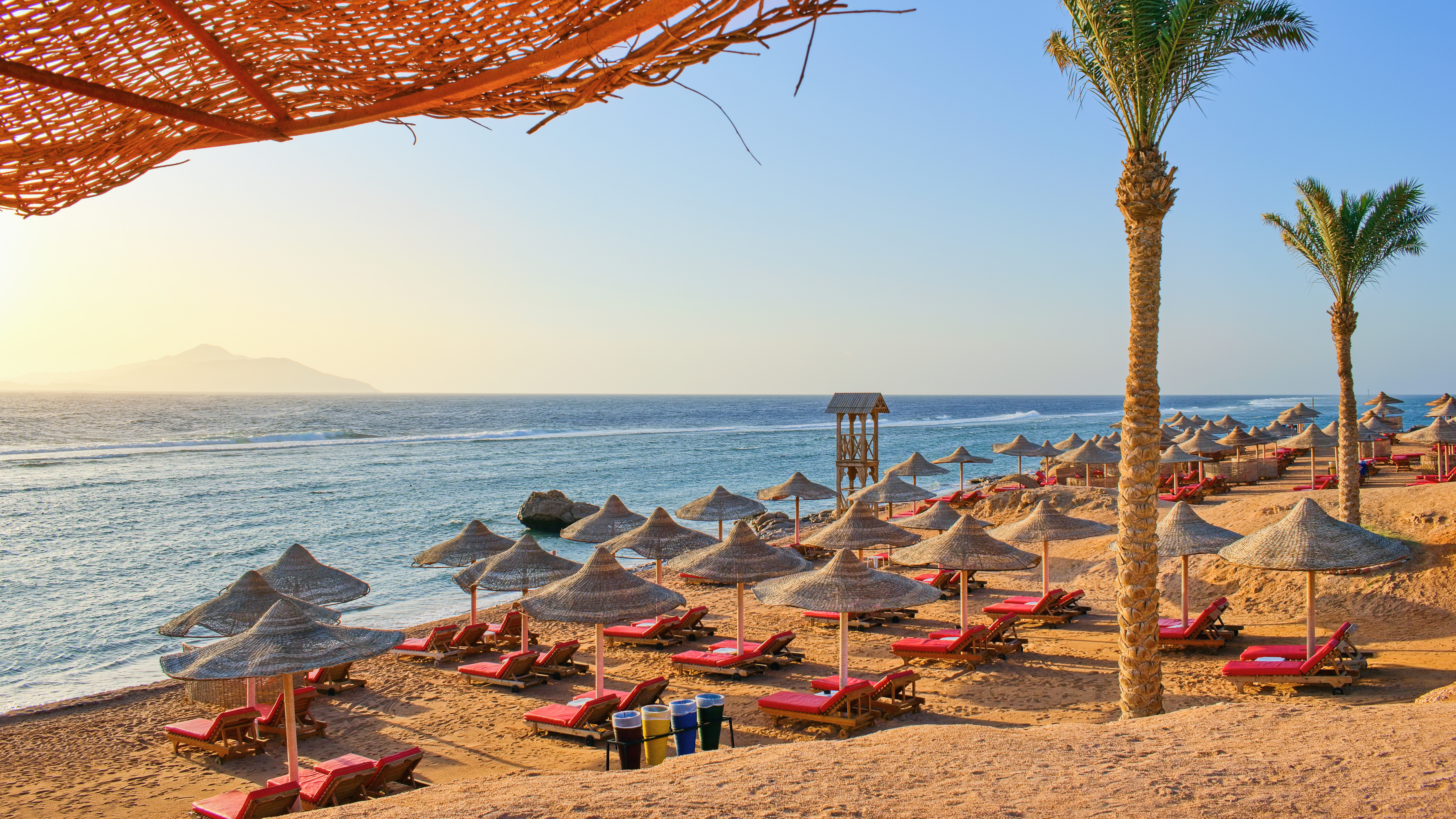 Rows of red lounge chairs with thatched straw umbrellas line a sunny, sandy beach overlooking the turquoise sea and distant mountains.