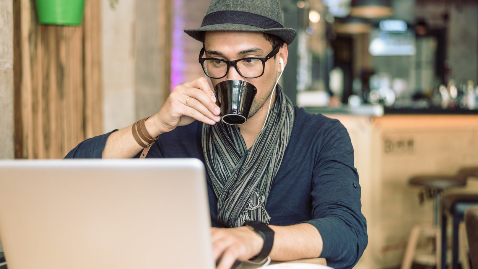 A man wearing a hat and scarf drinks from a cup while working on a laptop.