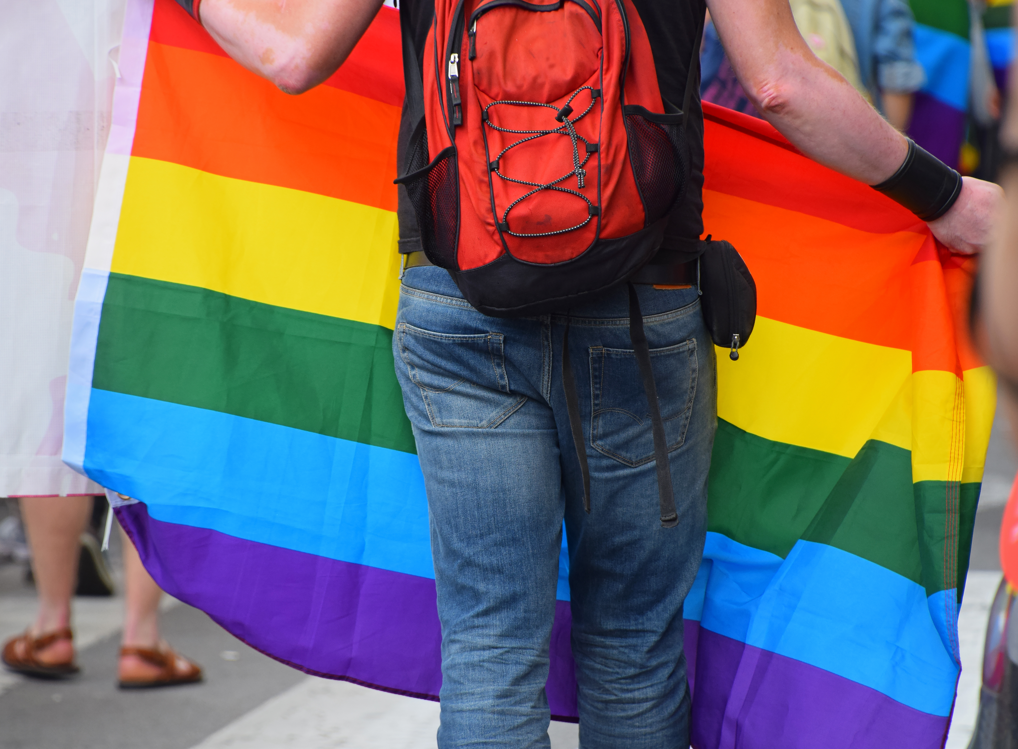 Person carrying a rainbow Pride flag seen from the back in jeans and with a red backpack