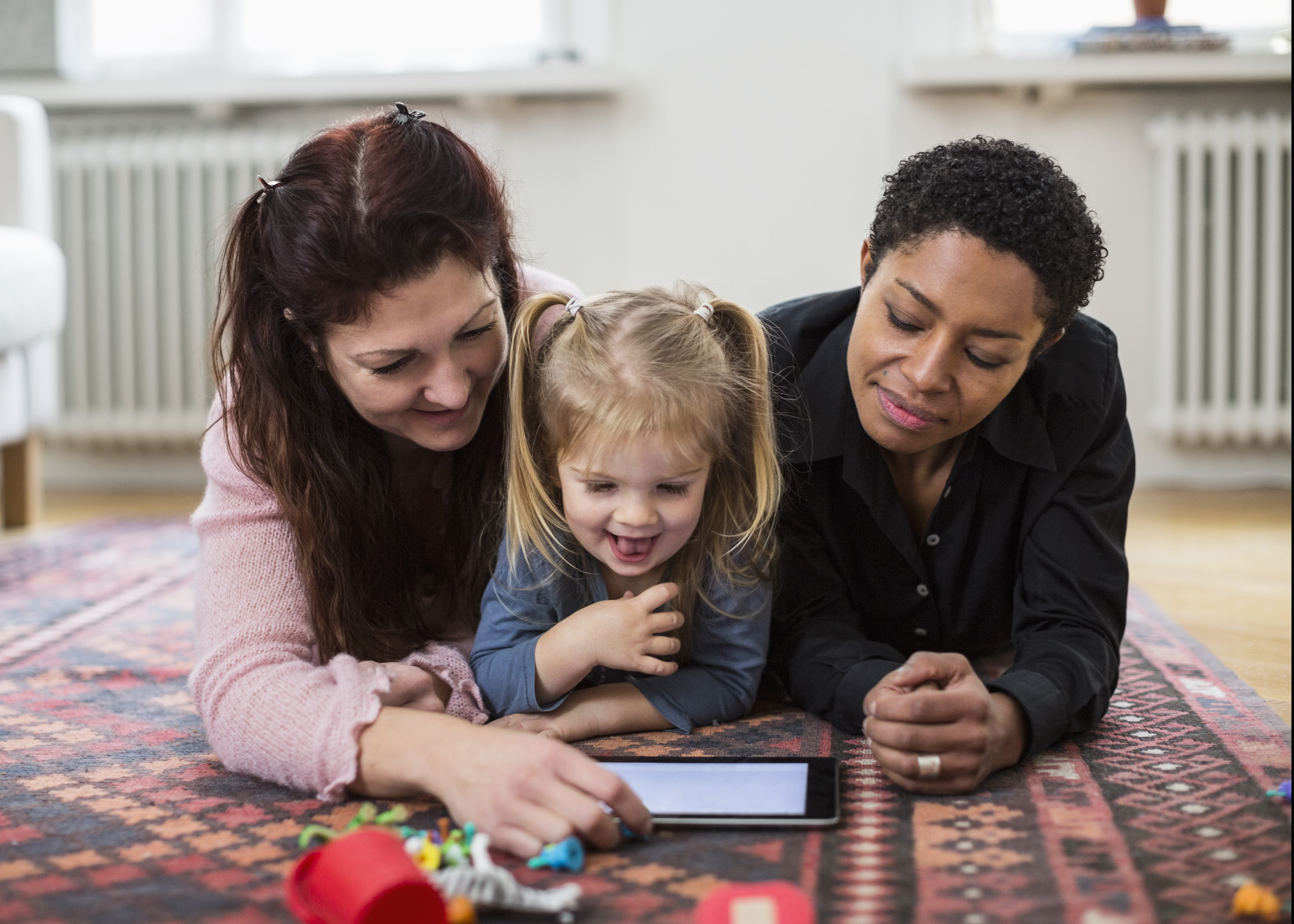Two women and a little girl playing together on a tablet while lying on the floor