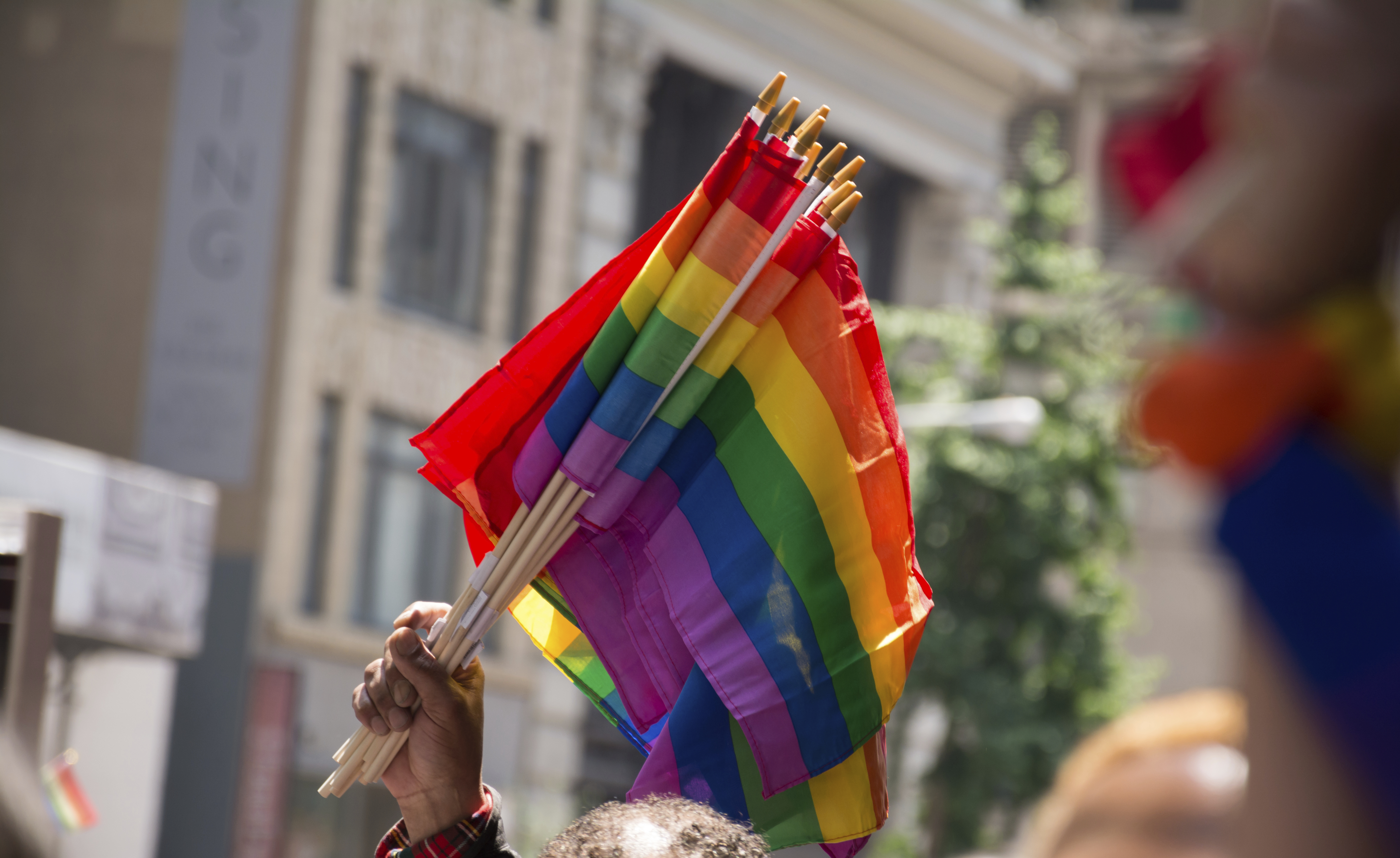 A hand holding up multiple rainbow colored flags in a city during a Pride parade